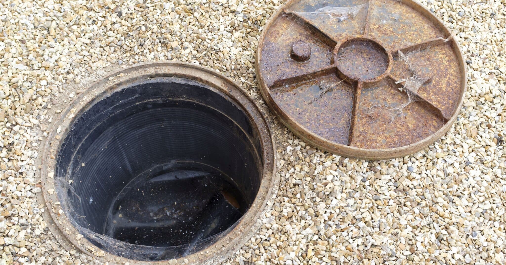 Chambre de visite ouverte dans une allée de gravier, couvercle en fonte déposé à côté : le regard de visite du raccordement à l’égout doit rester accessible